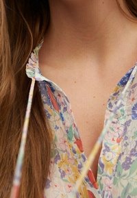 Blumenbluse mit Rüschenausschnitt, leichtes Material mit mehrfarbigen Blumen, Knopfverschluss, Bindebänder am Hals, weiche Textur.