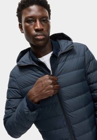 Giacca imbottita trapuntata blu navy, con chiusura a zip e cappuccio, caratterizzata da cuciture orizzontali e una consistenza morbida.