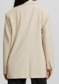 Beżowa marynarka o dopasowanym kroju, wykonana z gładkiego materiału, z długimi rękawami i rozcięciem z tyłu. Proste klapy bez ozdób.