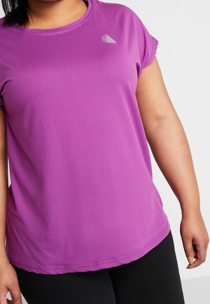 Kobieta w fioletowej sportowej koszulce z krótkim rękawem i małym białym logo przy dekolcie, ubrana w czarne spodnie na białym tle.