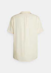 Camisa de manga corta en amarillo pálido, hecha de un tejido suave. Presenta un cuello botón y un solo pliegue en la parte trasera como detalle adicional.