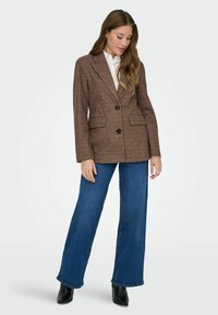 Bruine gestructureerde blazer met één knoop, twee frontzakken en gekartelde revers, gecombineerd met brede blauwe denim jeans en zwarte laarzen.