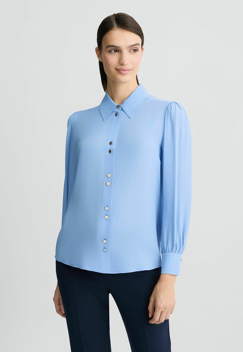 Lichtblauwe blouse met een spitse kraag, knoopsluiting aan de voorkant, lange pofmouwen en een gladde textuur. Bevat decoratieve knopen langs de split.
