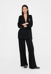 Blazer negro de doble botonadura combinado con pantalones negros de pierna ancha. El blazer presenta un escote profundo y un acento de broche plateado.