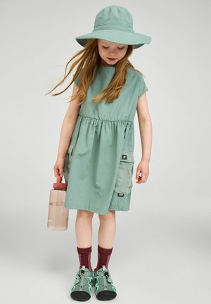 Młoda dziewczyna w zielonej sukience i kapeluszu z szerokim rondem trzyma butelkę z wodą, ma na sobie bordowe skarpetki i zielone trampki, stoi na prostym tle.