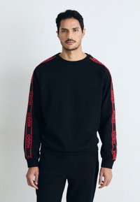 Svart hettegenser med røde logoaksenter på ermene, rund halsdesign, laget av teksturert stoff, vist i en nøytral bakgrunnsinnstilling.
