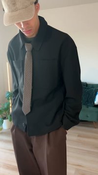 Uomo con berretto beige, giacca nera, cravatta marrone e pantaloni marroni in piedi all'interno vicino a un divano verde e una pianta in vaso.