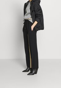 Svart vindjacka med huva, grå hoodie med guldlogotyp, svarta byxor med beige sidopaneler och svarta klackade ankelskor.