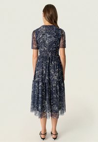 Žena v námořnickém modrém midi šatech s bílými vícestupňovými vzory, s průhlednými krátkými rukávy a černými páskovými podpatky, stojící zády k fotoaparátu.