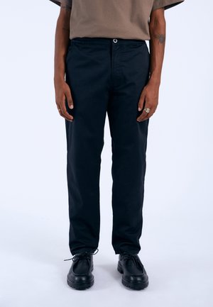 Pantaloni neri con gamba dritta, realizzati in un materiale liscio, dotati di chiusura con bottone e tasche. Indossati con scarpe nere con lacci.