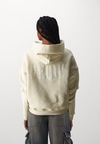 Krema barve kapucar z velikim vezenim napisom "PEQUIS" na hrbtu. Ima sproščen kroj, sprednji žep ter rebrast rob in manšete.