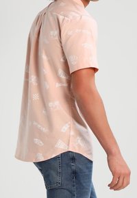 Kurzärmeliges, hellrosa Hemd mit weißen grafischen Drucken, darunter Logos und Designs. Der Stoff wirkt glatt mit einem lockeren Schnitt.