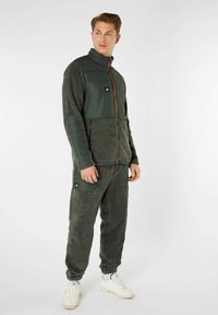 Giacca in pile verde con sezioni imbottite, chiusura con zip e tasche, abbinata a pantaloni in pile coordinati. Presenta un dettaglio con logo e polsini elastici.