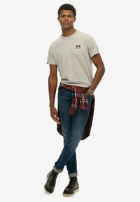 T-shirt grigio a maniche corte, jeans blu, stivali neri. Camicia a plaid rossa legata in vita. Logo notevole sulla tasca del petto della maglietta.