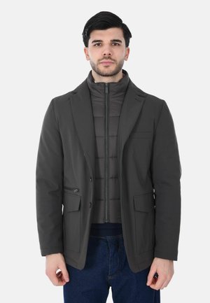 Cappotto grigio scuro con design a blazer strutturato, con chiusura frontale con zip e due tasche anteriori, indossato sopra una giacca imbottita.