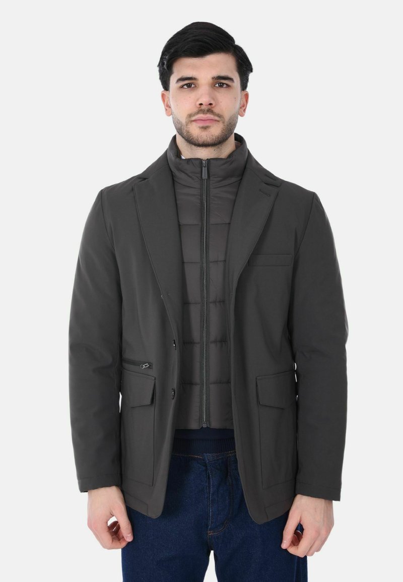 Cappotto grigio scuro con design a blazer strutturato, con chiusura frontale con zip e due tasche anteriori, indossato sopra una giacca imbottita.