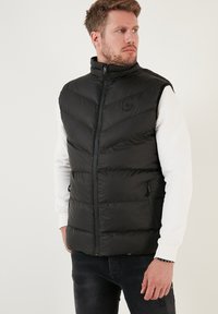 Crni puffa prsluk s visokim ovratnikom i dizajnom u obliku chevrona. Ima džepove sa zatvaračem i logotip na prsima. Glatka, podstavljena tekstura.