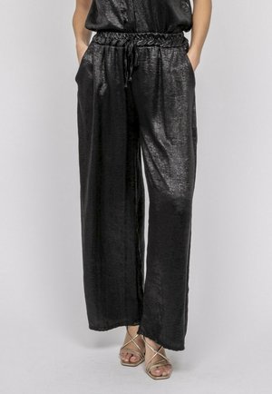 Femme portant un pantalon large en satin noir avec une taille élastique et un cordon de serrage, assorti à des sandales à talons hauts beige à brides.