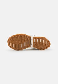 La semelle de chaussure Columbia présente un design en caoutchouc moulé avec des motifs hexagonaux, une couleur brun clair et deux bandes blanches d'accent.