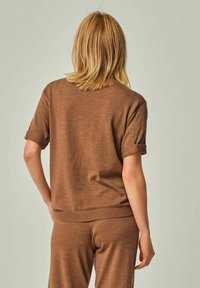 Woman with shoulder-length blonde hair wearing a brown short-sleeve knit top and matching pants, standing against a plain background.