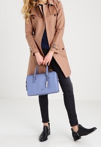 Brązowy płaszcz trench z granatową bluzką, czarnymi skinny jeansami i czarnymi botkami za kostkę; trzymając niebieską skórzaną torbę na ramię z srebrnymi akcentami.