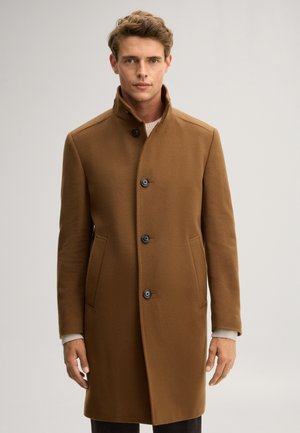 Mann mit kurzen braunen Haaren trägt einen geknöpften braunen Wollmantel und einen hellen Pullover, steht vor einem einfarbigen hellgrauen Hintergrund.