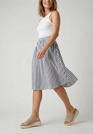 Frau geht und trägt ein weißes ärmelloses Oberteil, einen blau-weiß vertikal gestreiften Midirock und beige Slipper.