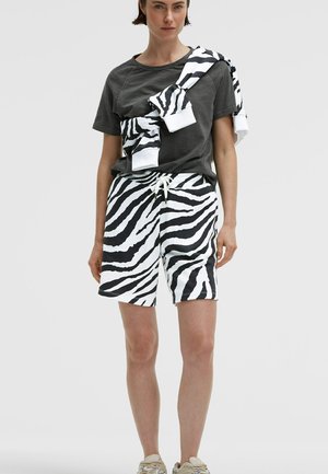 Frau trägt ein schwarzes T-Shirt und Shorts mit Zebra-Streifen, dazu ein passendes Sweatshirt mit Zebra-Streifen, das über der Schulter gebunden ist, vor einem schlichten Hintergrund stehend.