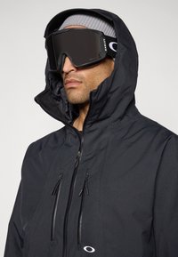 Man wearing black waterproof hooded jacket, gray beanie, and large black Oakley ski goggles against plain background.