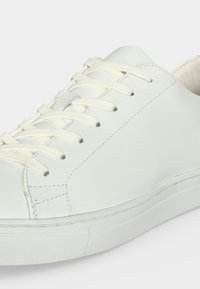 Baskets basses blanches avec lacets blancs, dessus en cuir lisse et semelle en caoutchouc sur un fond clair uni.