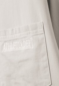 Close-up of beige fabric pocket with stitched "ATHLETIX DEPT" logo in matching thread on garment.