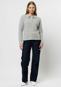 Žena oblečená v svetlosivej pletenej svetri s golierom, tmavomodrých džínsach a bielych teniskách, stojaca pred jednoliatym bielym pozadím.