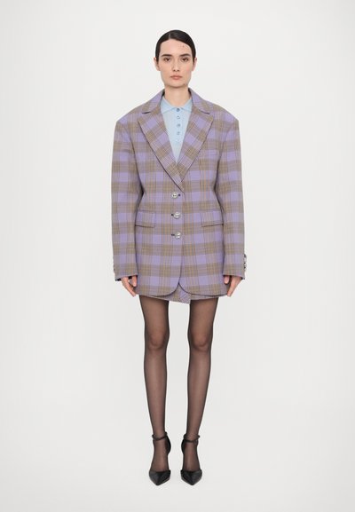 Kvinna står klädd i en överdimensionerad lila rutiga blazer och matchande shorts, ljusblå skjortblus, svarta genomskinliga strumpbyxor och svarta klackskor.