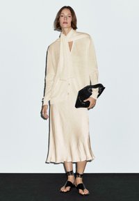 Beige cotton cardigan over a cream satin dress, paired with black strappy sandals and a black textured clutch. Smooth finish, relaxed fit.