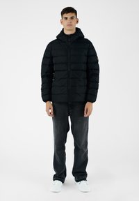 Giacca a vento nera con zip frontale, cappuccio e polsini a costine. Indossata con jeans scuri e sneakers bianche su uno sfondo semplice.