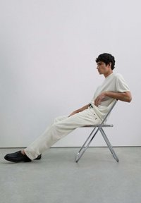 Modelo sentado en una silla metálica plegable, vistiendo una camiseta de punto beige claro, pantalones color crema y zapatos negros. Estética simple y minimalista.