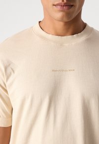 Beige cotton t-shirt with classic crew neck, short sleeves, and subtle "Marc O'Polo DENIM" logo in gold on the front. Smooth texture.