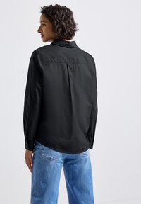 Donna con capelli ricci corti, che indossa una maglietta nera a maniche lunghe e jeans azzurri chiari, in piedi di spalle contro uno sfondo neutro.