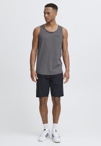 Graues ärmelloses Tanktop mit einer Brusttasche, kombiniert mit schwarzen Shorts. Das Modell trägt weiße Sportschuhe mit schwarzen Streifen und weiße Socken.