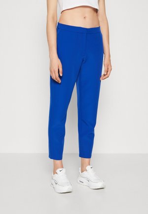 Vrouw met felblauwe cropped broek en witte sneakers, staand tegen een effen lichte achtergrond.