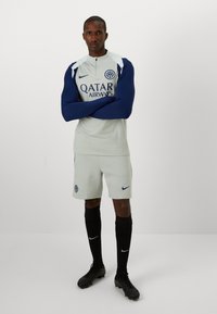 Witte sporttrui met marineblauwe mouwen, voorzien van een ritssluiting en branding. Gekleed met grijze shorts en zwarte kniekousen, zwarte voetbalschoenen.