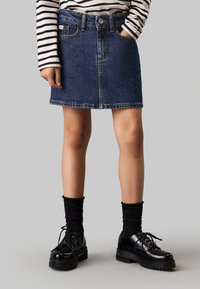 Gonna di jeans di colore blu scuro, sopra il ginocchio, con design a cinque tasche, passanti per cintura e orlo sfilacciato. Abbinata a scarpe nere con lacci spessi e calze.