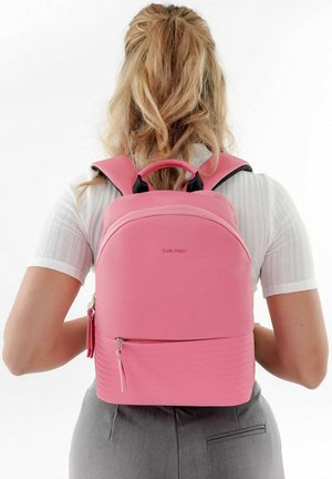 Sac à dos en tissu rose avec une surface texturée, deux bretelles, une poignée supérieure et une poche zippée à l'avant. Le logo de la marque est visible.