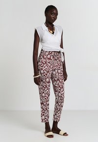 Donna in piedi con pantaloni a motivi bordeaux e bianchi, blusa senza maniche bianca a vestaglia, sandali beige e collana di perline spesse su uno sfondo neutro.