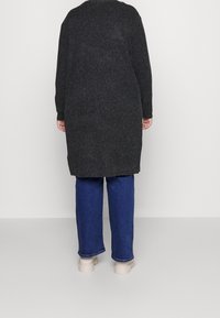 Žebrovaný kardigan z uhlíkové příze s dlouhými rukávy a rozšířeným spodním lemem, v kombinaci s modrými džíny a béžovými botami, zobrazen zezadu na neutrálním pozadí.