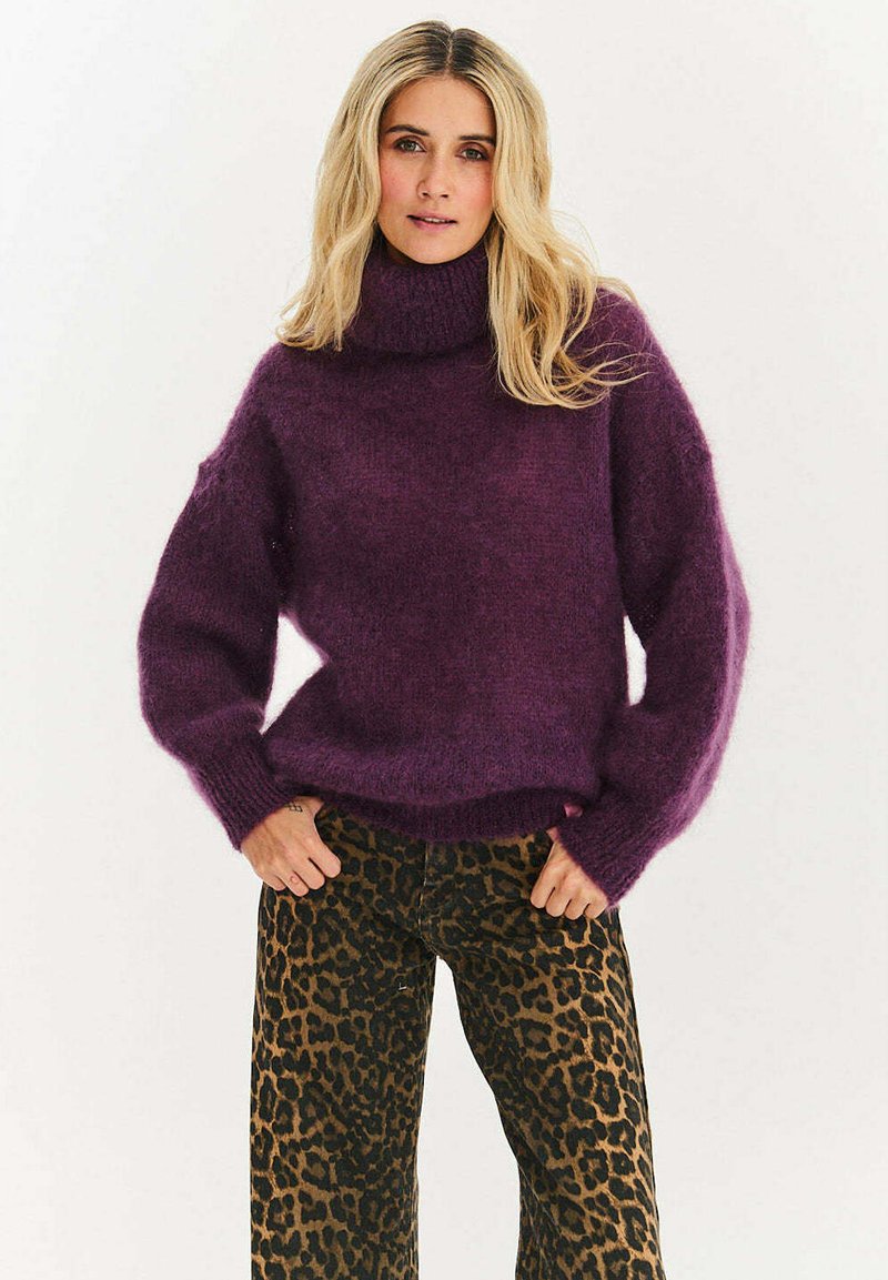 Žena s dlhými blond vlasmi, oblečená v hrubom fialovom roláku a nohaviciach so vzorom leopardej srsti, stojaca s rukami v vreckách.