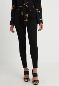 Mujer vestida con jeans ajustados de tiro alto negros, blusa de manga larga con estampado floral atada, y sandalias de tacón bloque abiertas en negro, sobre fondo blanco.