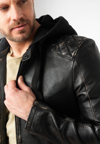 Schwarze Lederjacke mit gesteppten Schulterakzenten und einer schwarzen Kapuze. Der Reißverschluss und die Nähte sind sichtbar und zeigen eine strukturierte Oberfläche.