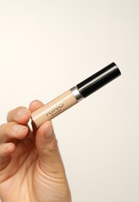 KIKO Milano concealer in een heldere tube, beige tint, met een zwarte dop en zilveren accent. Tussen de vingers gehouden tegen een neutrale achtergrond.