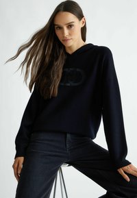 Felpa blu marina con un logo testurizzato al centro, abbinata a jeans scuri. Il tessuto appare morbido con una vestibilità comoda e maniche lunghe.
